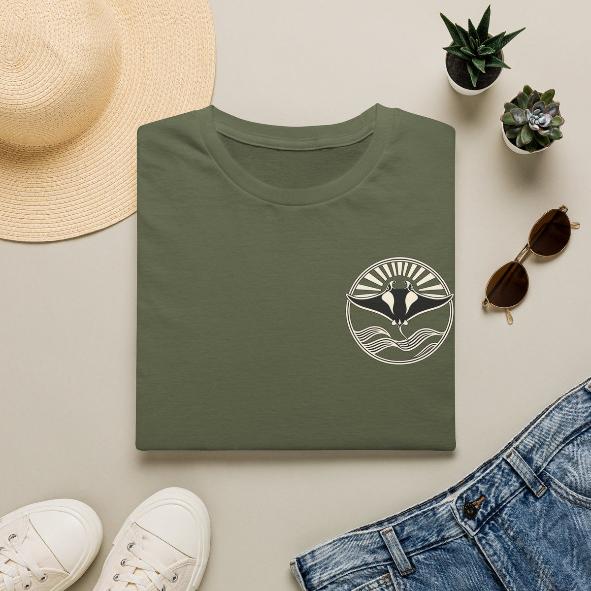 Folded military green t-shirt showing the small manta ray sunrise badge on the chest, flat lay with sunglasses and jeans