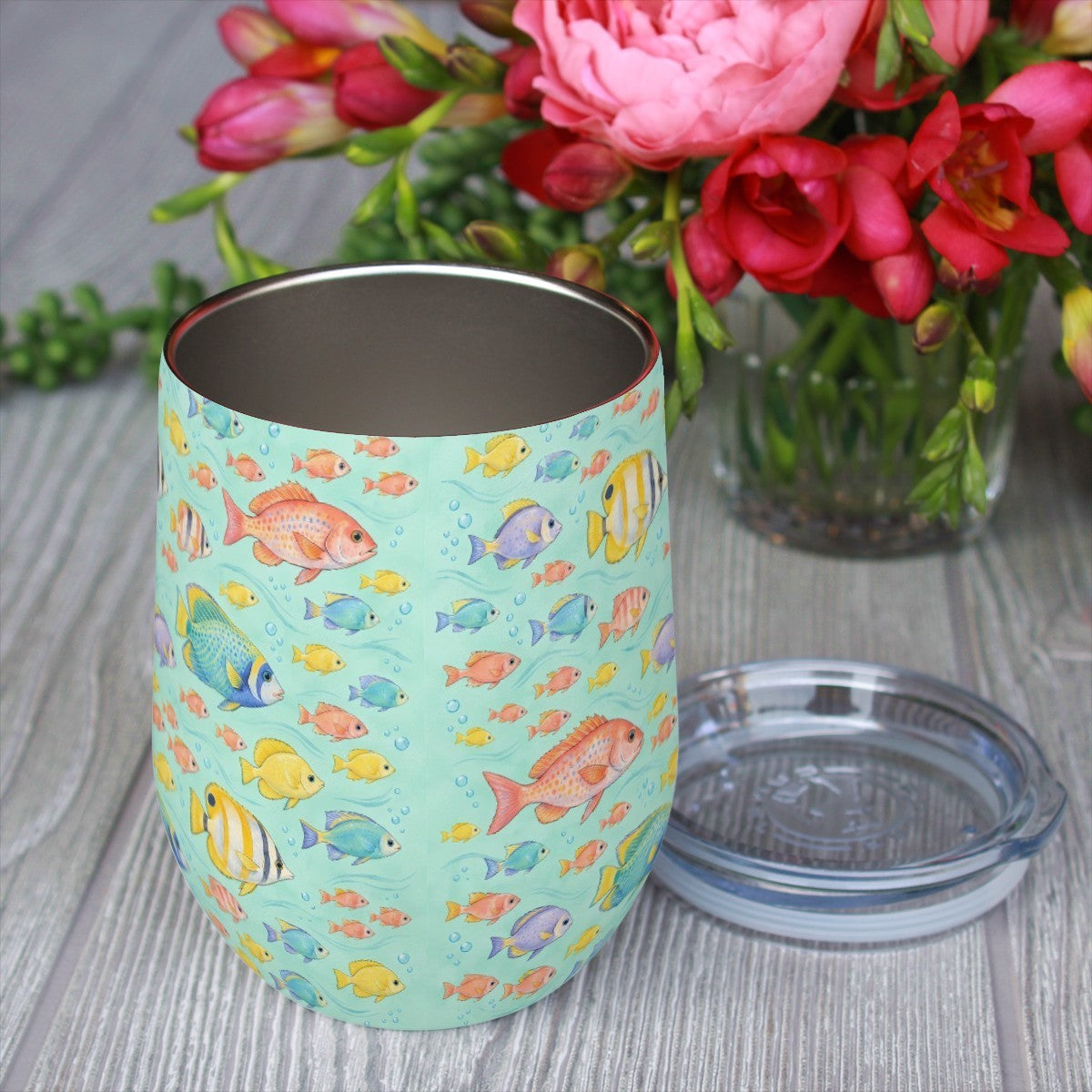 Reef fish pattern tumbler with its clear lid beside it on a wooden table, red flowers in the background