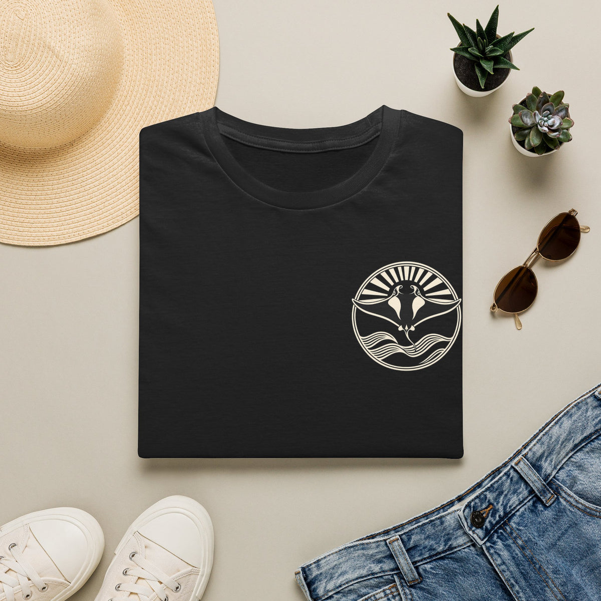 Folded black t-shirt showing the small manta ray sunrise badge on the chest, flat lay with sunglasses and jeans