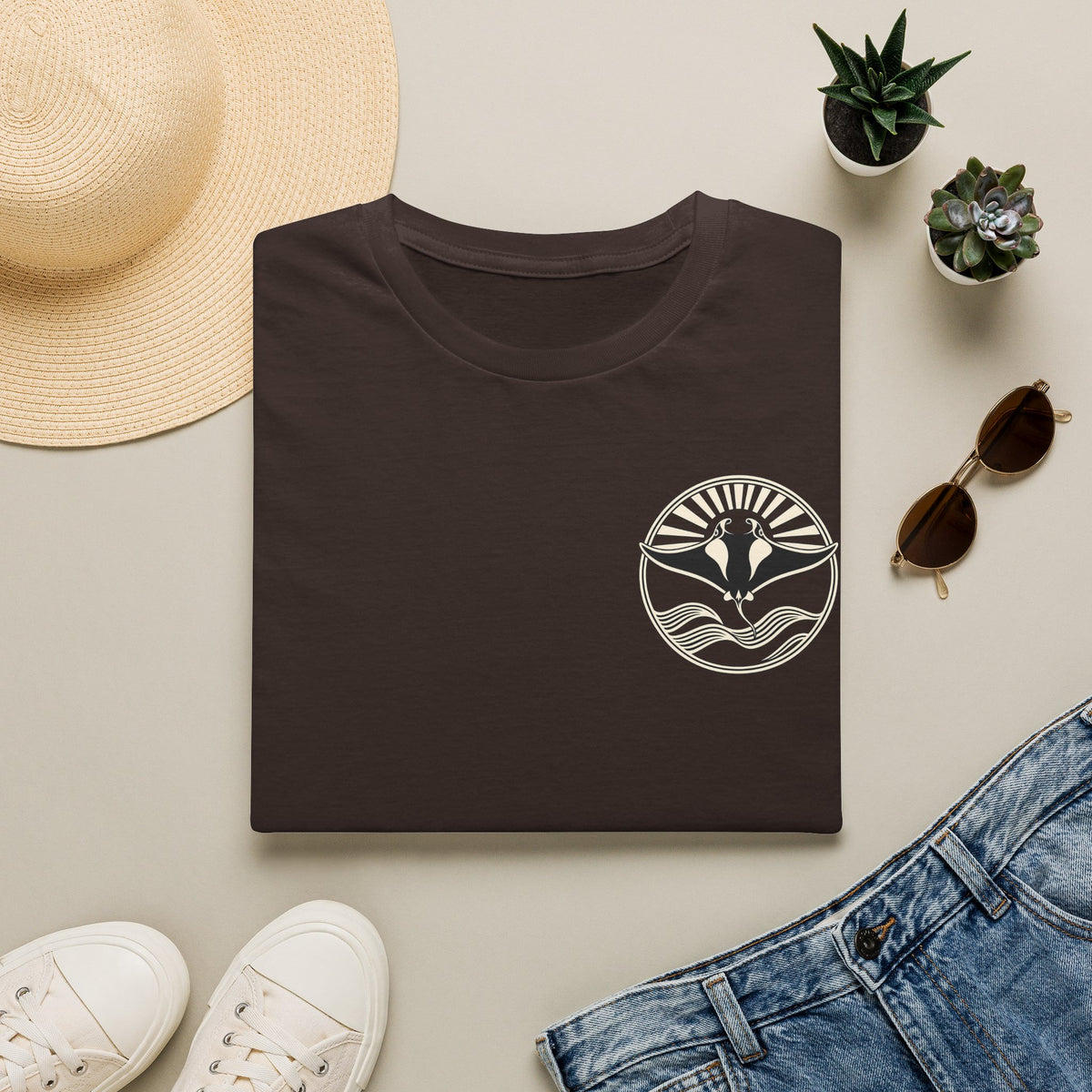 Folded dark chocolate t-shirt showing the small manta ray sunrise badge on the chest, flat lay with sunglasses and jeans