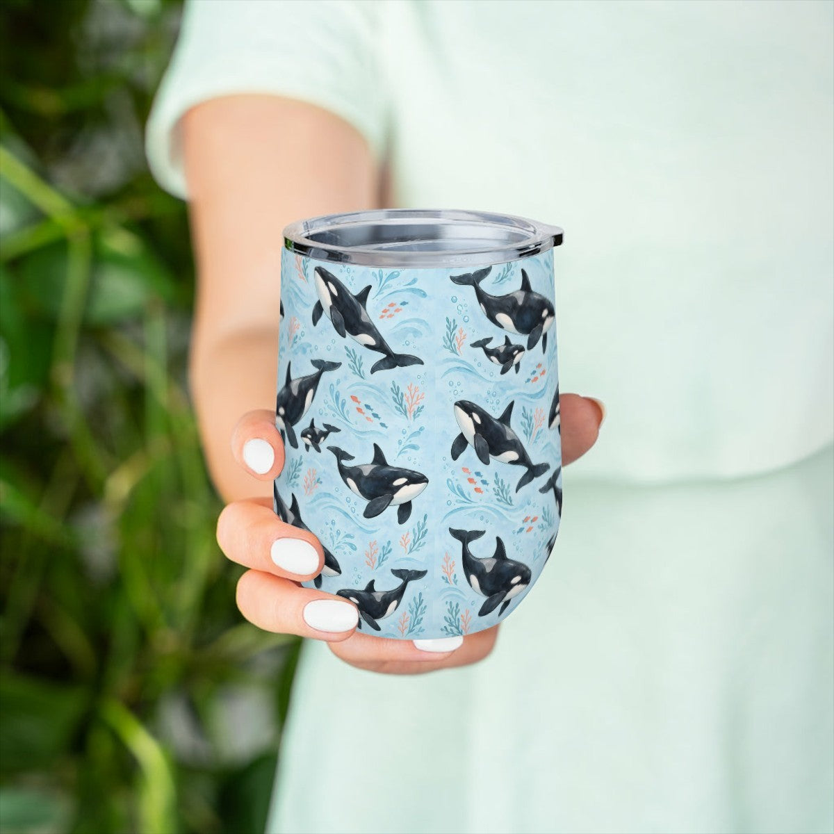 Hand holding the orca pattern tumbler in front of green leaves, with a pale green top behind