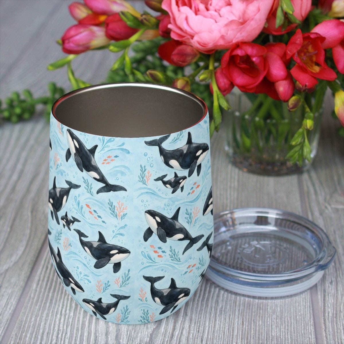 Orca pattern tumbler with its clear lid beside it on a wooden table, red flowers in the background