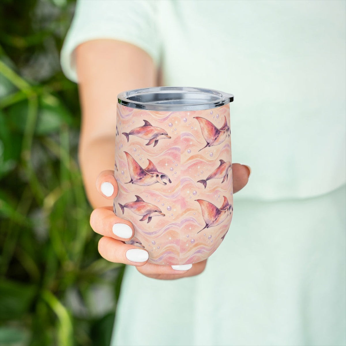Hand holding the dolphin and ray pattern tumbler in front of green leaves, with a pale green top behind