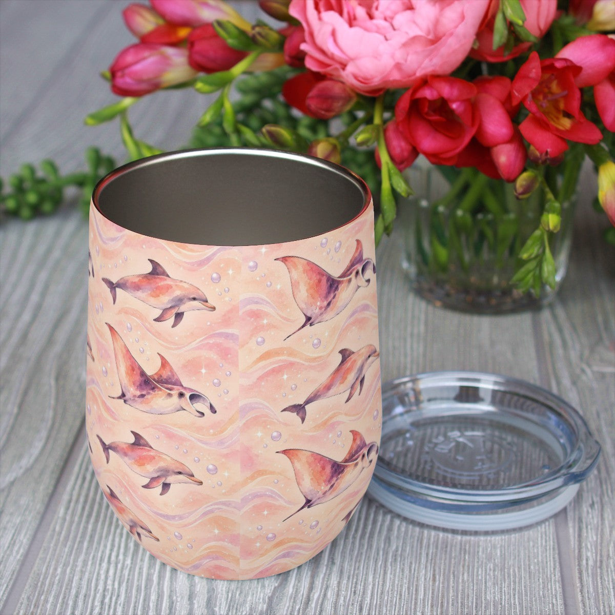 Dolphin and ray pattern tumbler with its clear lid beside it on a wooden table, red flowers in the background