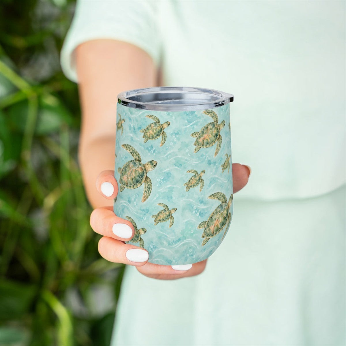 Hand holding the sea turtle pattern tumbler in front of green leaves, with a pale green top behind