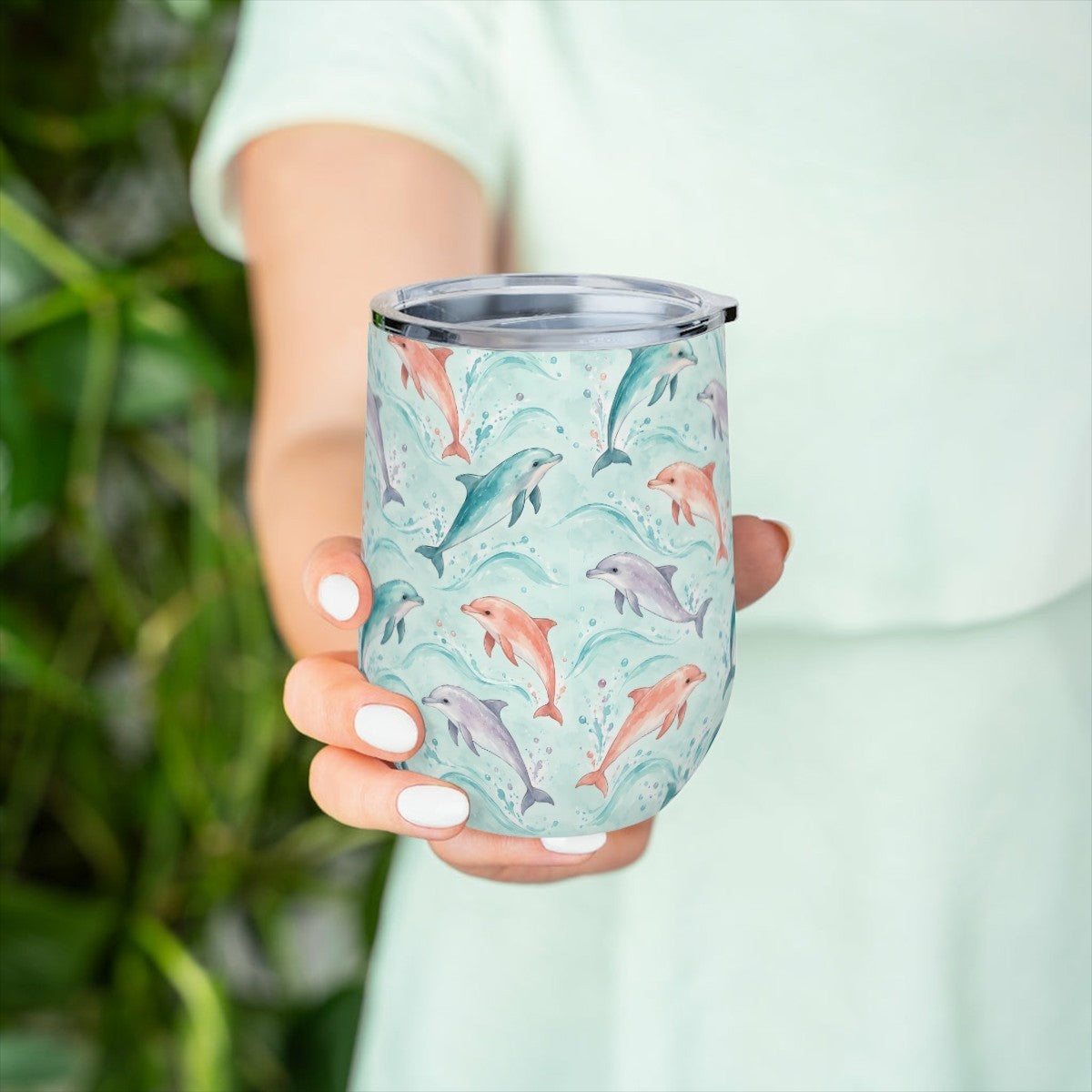 Hand holding the dolphin pattern tumbler in front of green leaves, with a pale green top behind