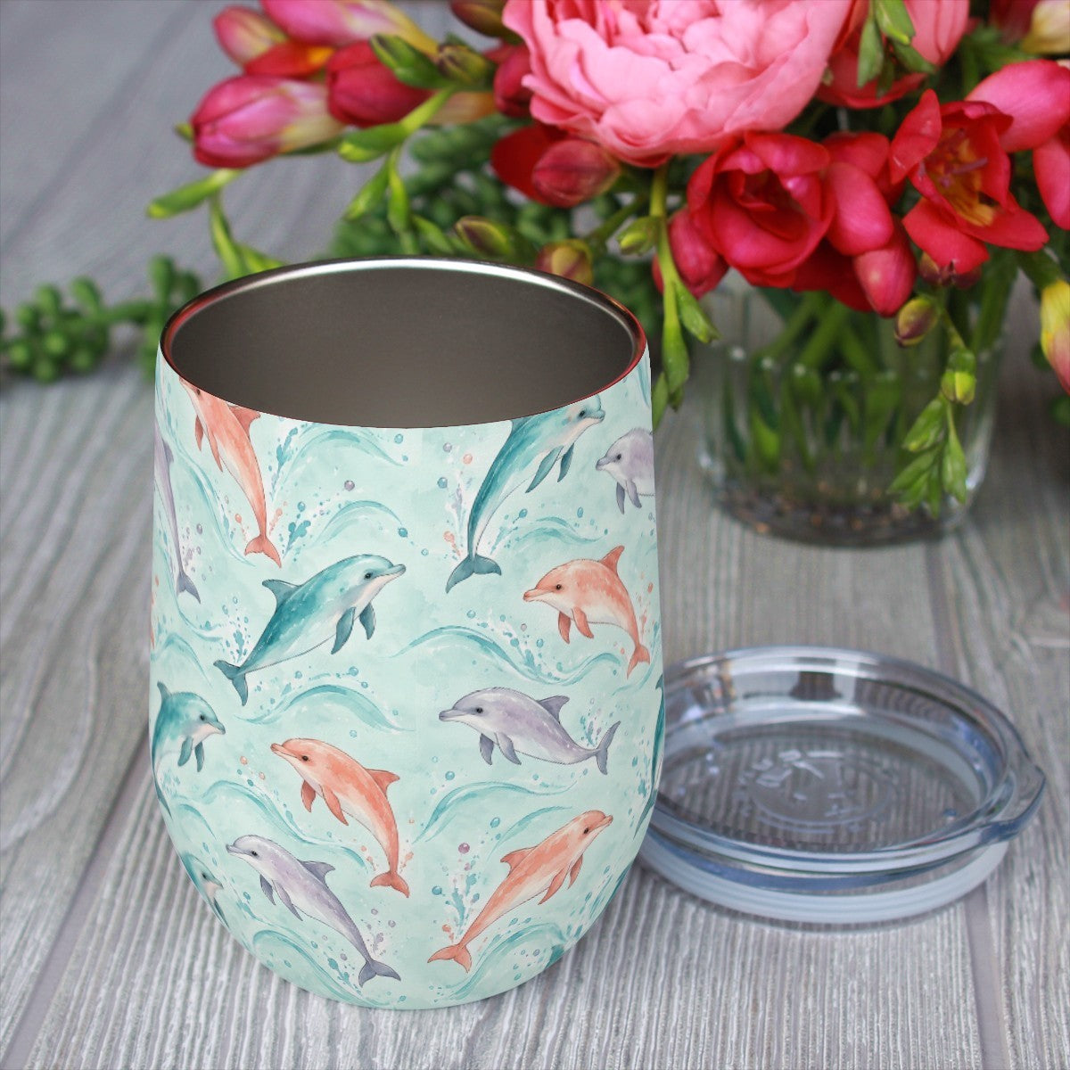 Dolphin pattern tumbler with its clear lid beside it on a wooden table, red flowers in the background