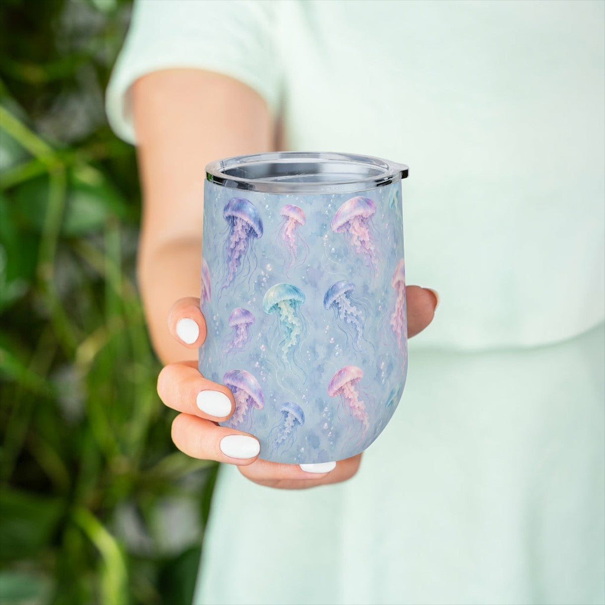 Hand holding the jellyfish pattern tumbler in front of green leaves, with a pale green top behind