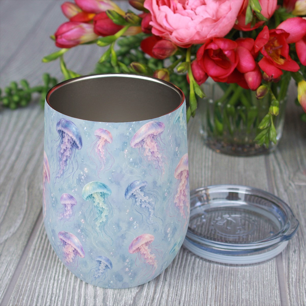 Jellyfish pattern tumbler with its clear lid beside it on a wooden table, red flowers in the background