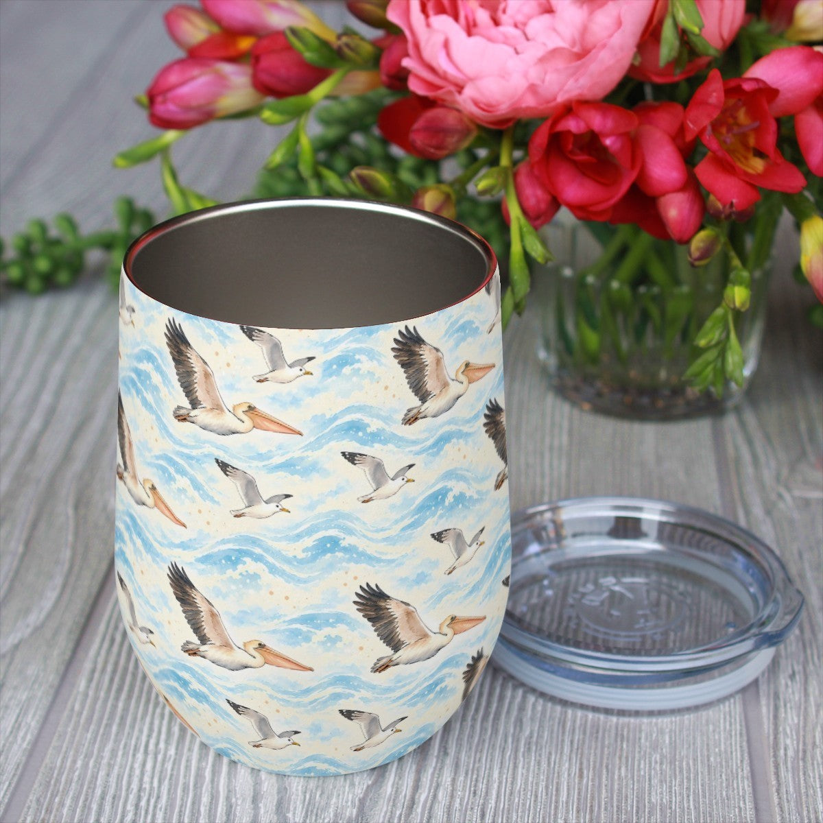 Pelican pattern tumbler with its clear lid beside it on a wooden table, red flowers in the background