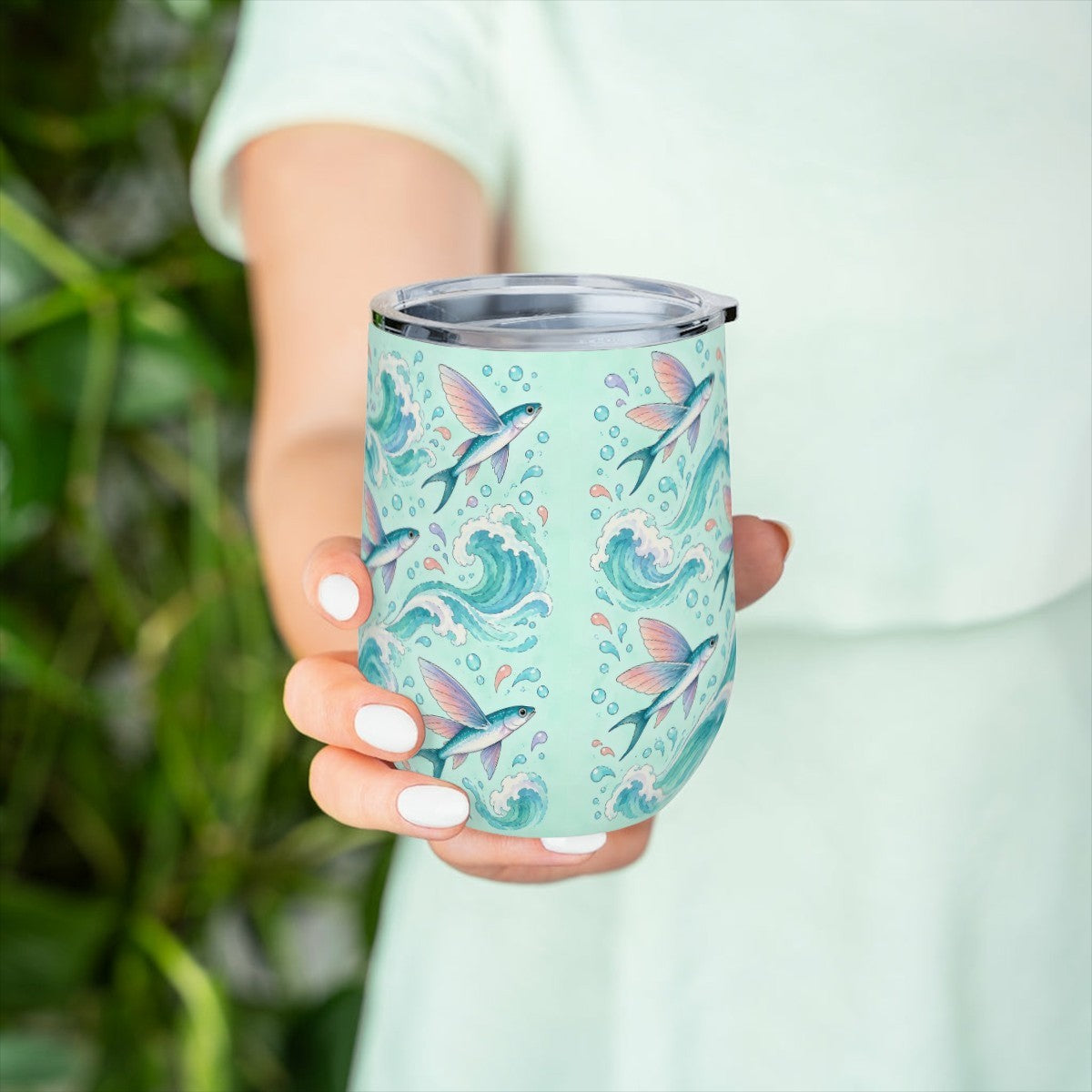 Hand holding the flying fish pattern tumbler in front of green leaves, with a pale green top behind