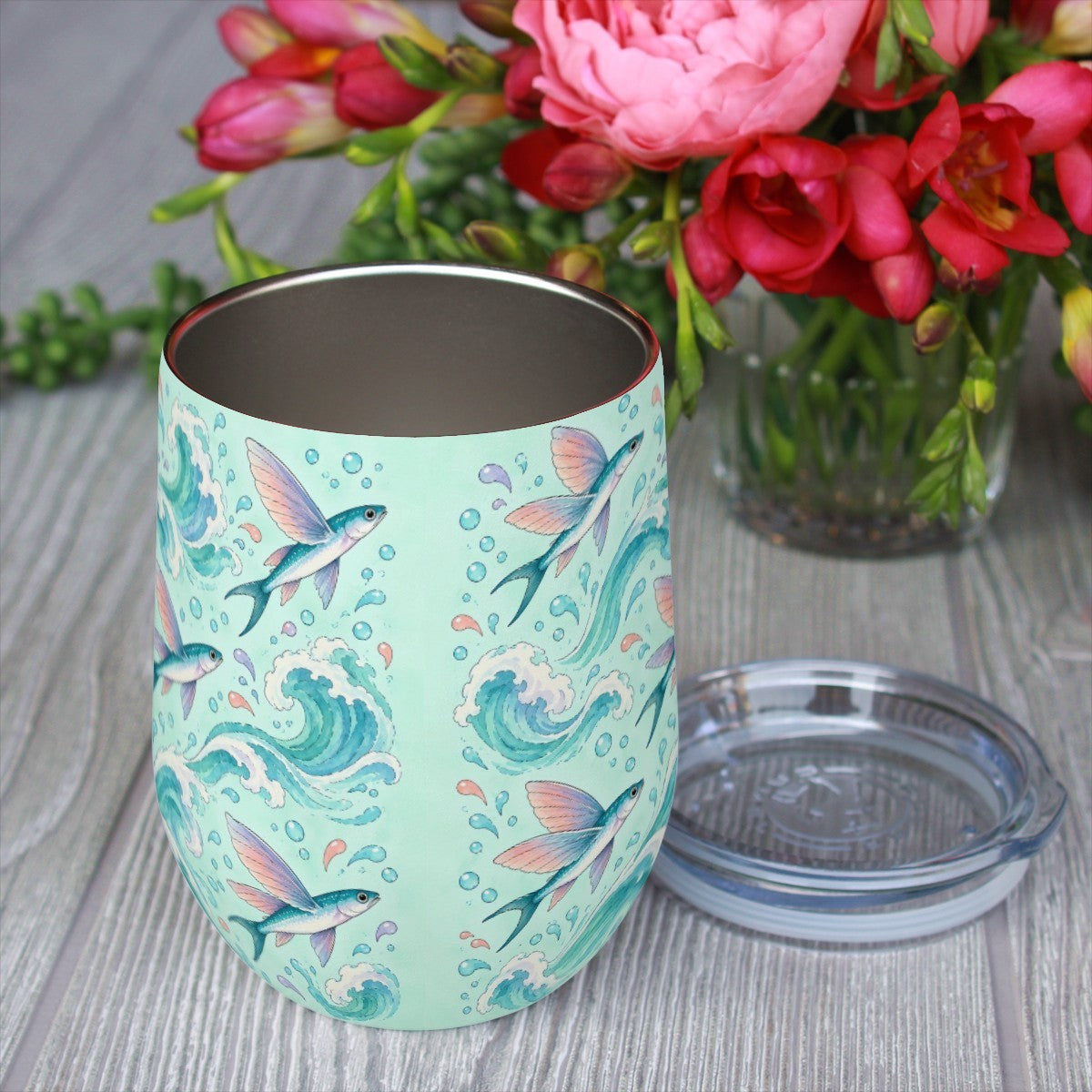 Flying fish pattern tumbler with its clear lid beside it on a wooden table, red flowers in the background