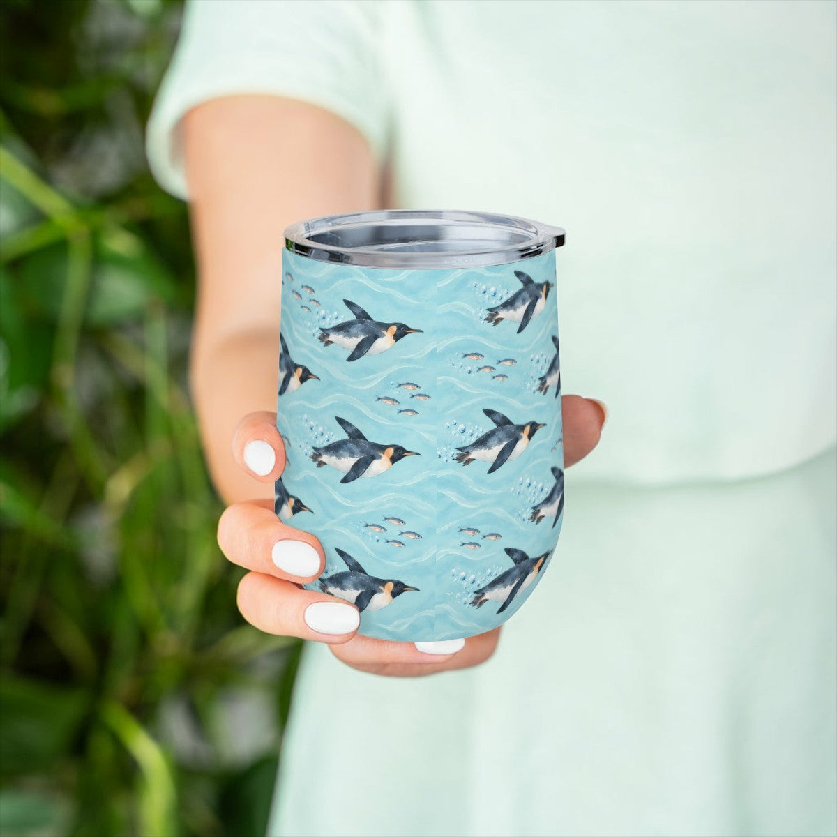 Hand holding the penguin pattern tumbler in front of green leaves, with a pale green top behind