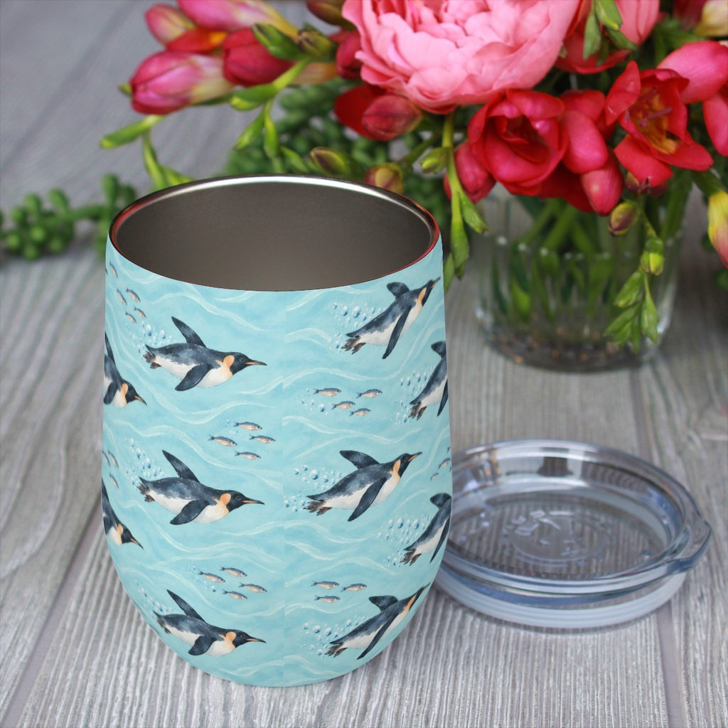Penguin pattern tumbler with its clear lid beside it on a wooden table, red flowers in the background