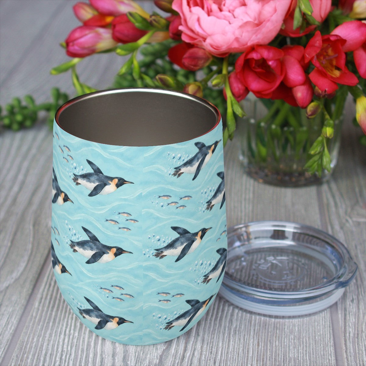 Penguin pattern tumbler with its clear lid beside it on a wooden table, red flowers in the background