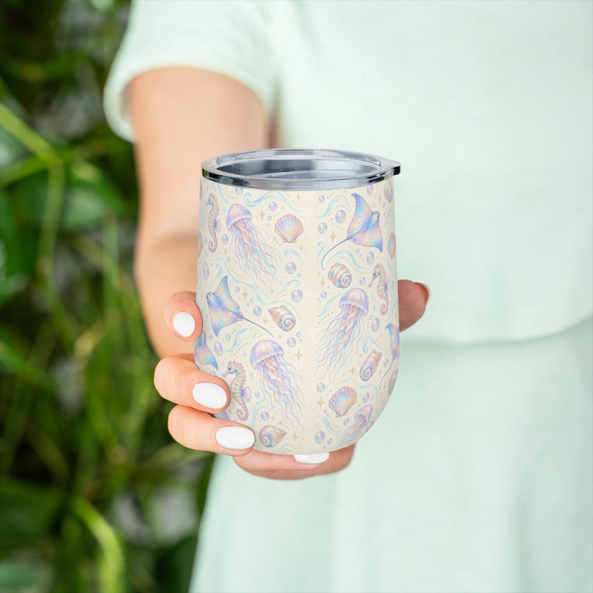 Hand holding the jellyfish and seahorse pattern tumbler in front of green leaves, with a pale green top behind