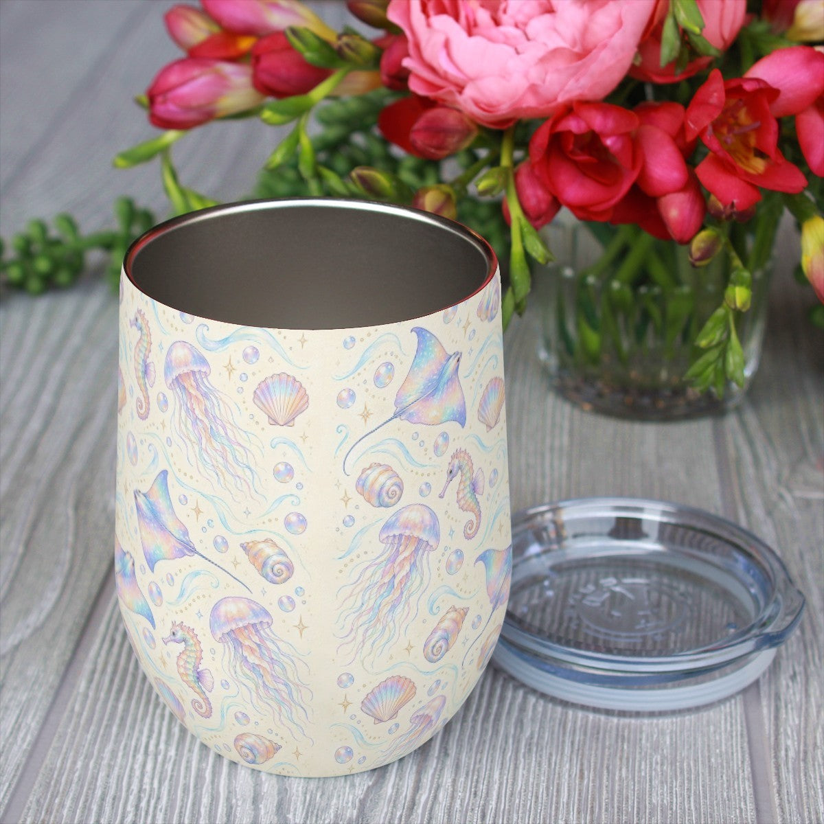 Jellyfish and seahorse pattern tumbler with its clear lid beside it on a wooden table, red flowers in the background