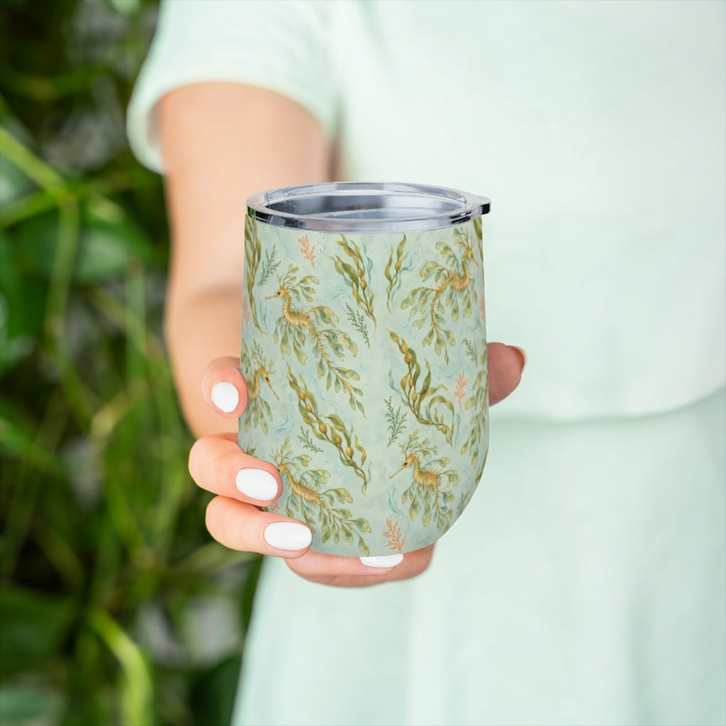 Hand holding the leafy sea dragon pattern tumbler in front of green leaves, with a pale green top behind