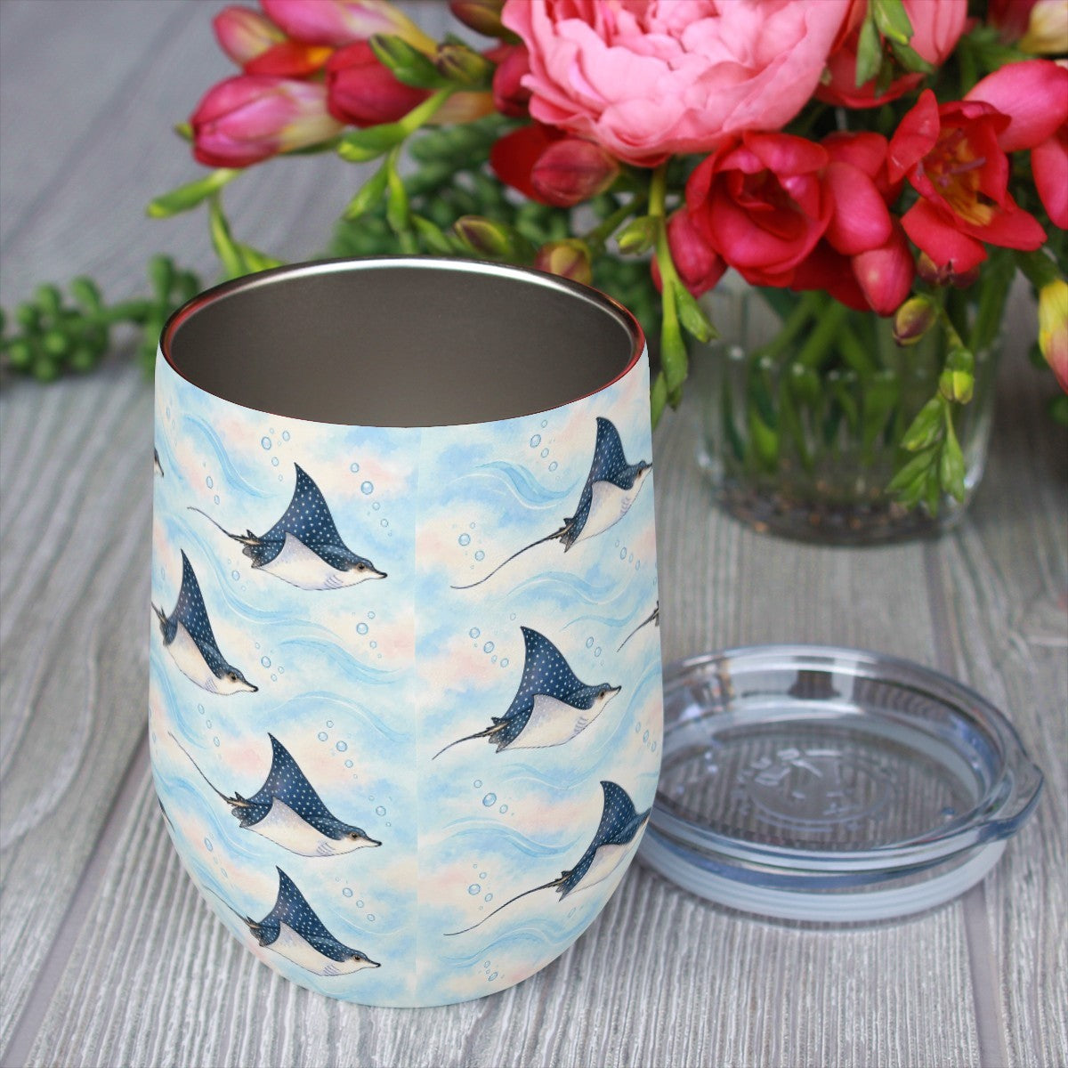 Eagle ray pattern tumbler with its clear lid beside it on a wooden table, red flowers in the background