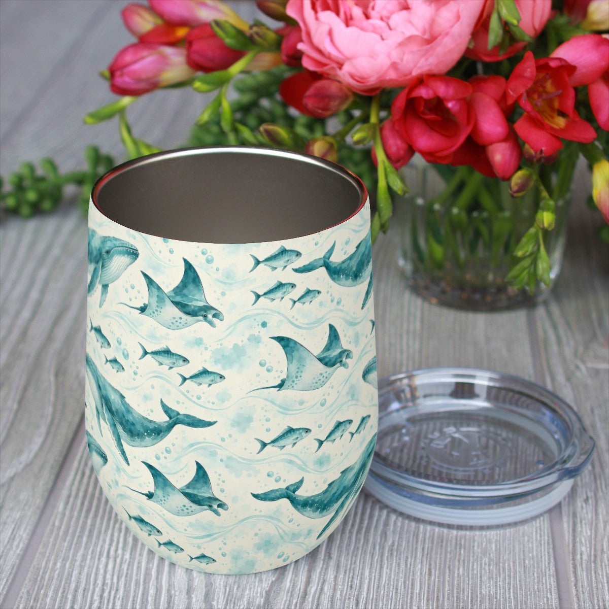Whale and ray pattern tumbler with its clear lid beside it on a wooden table, red flowers in the background