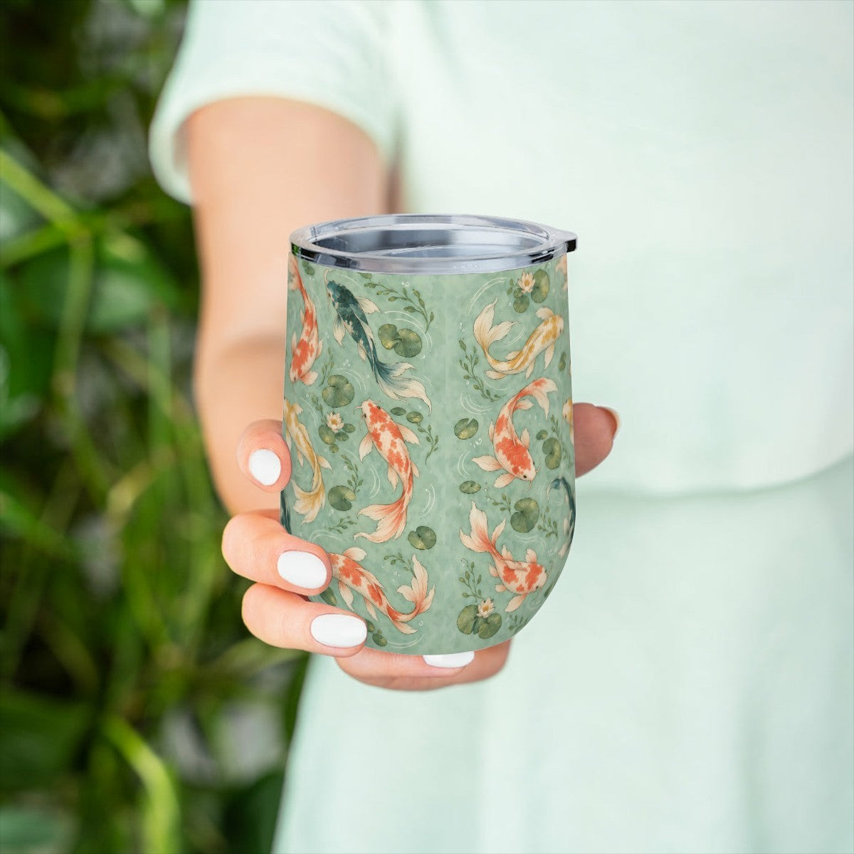 Hand holding the koi pattern tumbler in front of green leaves, with a pale green top behind