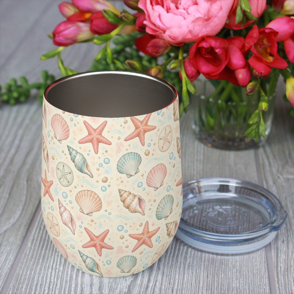 Seashell pattern tumbler with its clear lid beside it on a wooden table, red flowers in the background