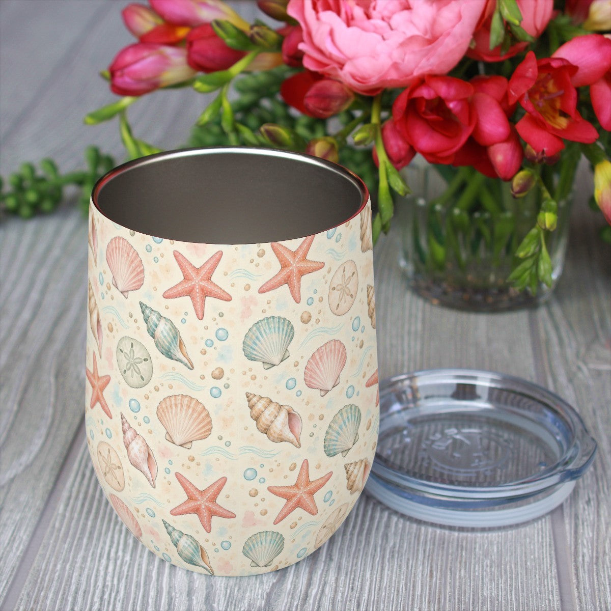 Seashell pattern tumbler with its clear lid beside it on a wooden table, red flowers in the background
